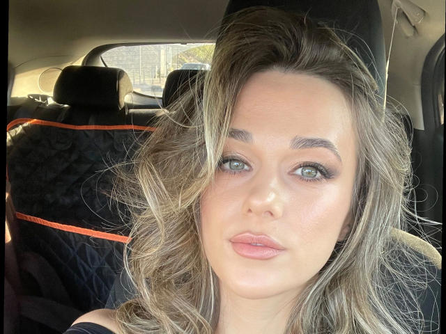 Minnie-hot with wavy blonde hair, green eyes, and pink lips, sitting in a car, wearing a black seat with orange trim, looking at the camera with a neutral expression.