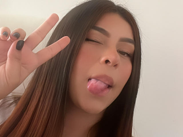 GabrielaFerrer with long brown hair, winking, tongue out, making a peace sign with black-painted nails, showing off her cute face and subtle makeup.