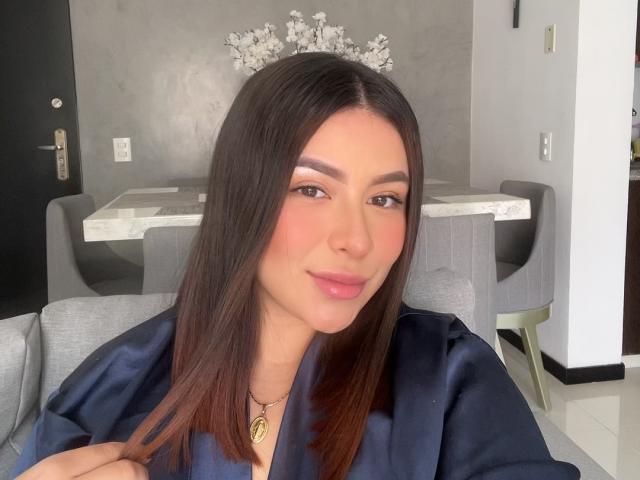 GabrielaFerrer with long brown hair, wearing a black satin robe, and a silver tiara, has pink lips, brown eyes, and a necklace with a gold pendant.