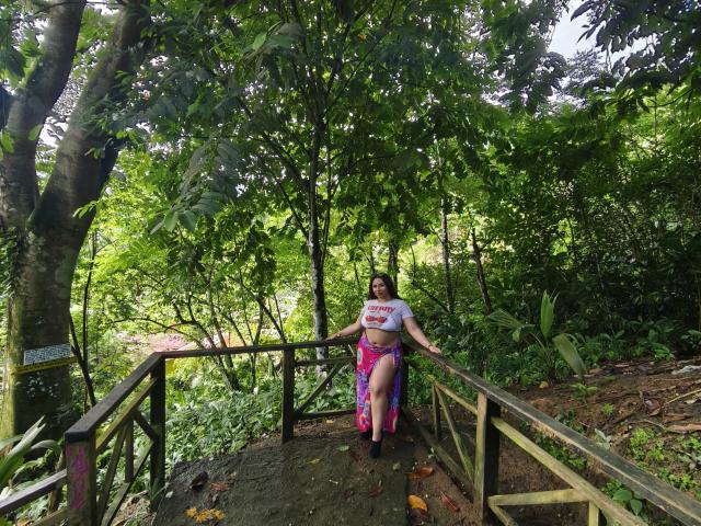 GabrielaFerrer stands on a wooden bridge, wearing a white crop top and pink, purple, and blue skirt, showing her ass through a high slit. Green forest background.