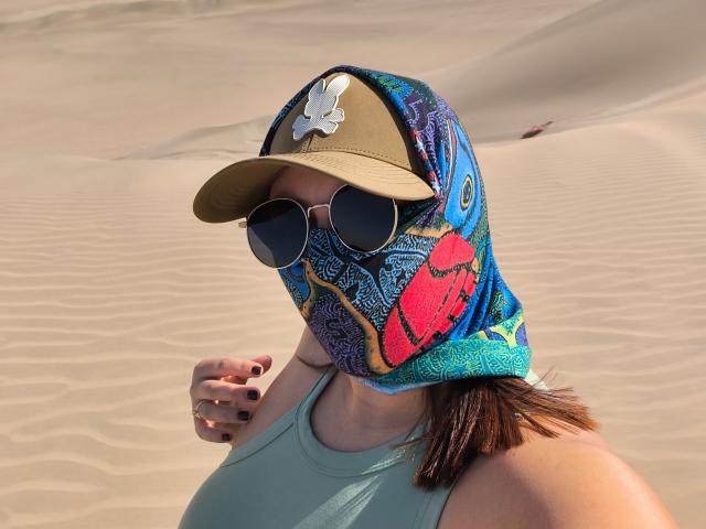 GabrielaFerrer with tan cap, colorful mask, round sunglasses, and teal tank top, showing her tits and hand with black nails, in a sandy desert.