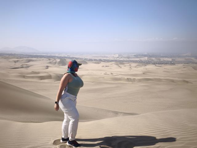 GabrielaFerrer with a blue and red headscarf, gray sleeveless top, white pants, black shoes, standing in a sandy desert, shadow on the sand, clear blue sky.