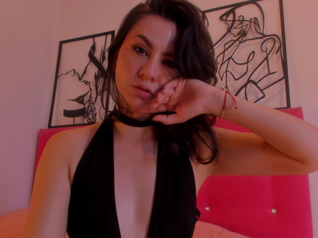 Kari Carter mit dunklem Haar, schwarzem Choker und einem tief ausgeschnittenen schwarzen Oberteil, die sich ihr Gesicht berührt, wobei ihre kleinen Brüste sichtbar sind, ein rosafarbenes Schlafzimmer im Hintergrund, ein rotes Armband und zwei abstrakte Wandbilder..
