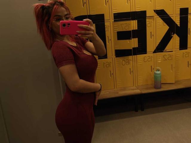 MarianneBowe with red hair in pigtails, wearing glasses, red tight dress, holding pink phone, showing curvy butt and small tits, standing in locker room, water bottle on bench.