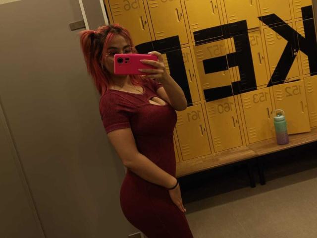 MarianneBowe with red hair in a tight maroon dress, showing her curvy ass and small tits, taking a mirror selfie in a locker room, holding a pink phone.