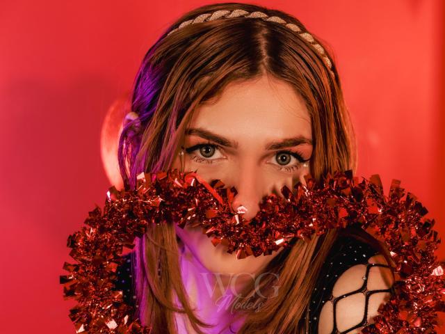 AndyDupont with brown hair, blue eyes, and a braided headband, wearing a red tinsel garland covering her mouth, against a red background. Watermark "W&N" visible.
