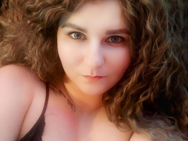 MinistraX has curly brown hair, light skin, and blue eyes, wearing a black lace top, with a tattoo on her left shoulder, looking directly at the camera.