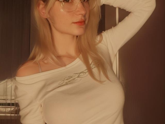 MiraliXSweet with blonde hair, glasses, and off-shoulder white top, showing her tits, arm raised, in a warm, softly-lit image.