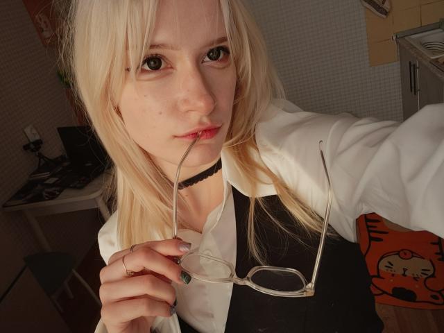 MiraliXSweet with blonde hair, holding glasses in front of her slightly parted lips, wearing a white shirt and black choker, in a dimly lit room.