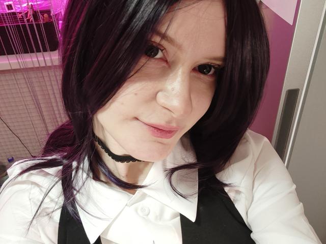 MiraliXSweet has pale skin, black hair with purple tips, and is wearing a white shirt with a black choker and black vest. She's smiling at the camera.