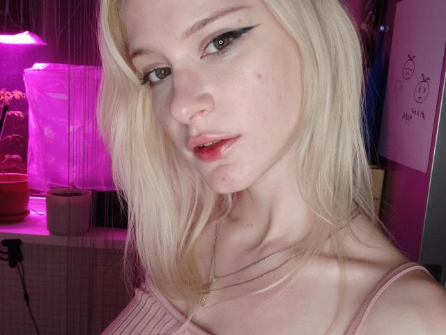 Close-up of MiraliXSweet, a blonde woman with light skin and black eyeliner. She has slightly parted pink lips and a small pimple on her right cheek. Her hair is straight and falls loosely around her face. She wears a pink, ribbed tank top and a thin gold necklace with a small cross pendant. The pink and purple lighting in the background adds a subtle, warm glow to her skin. The image focuses on her face and upper chest. She has a slight, confident expression.
