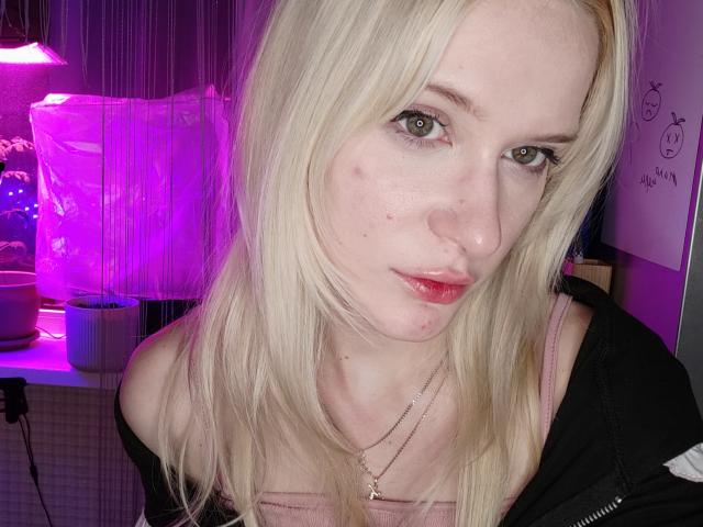 MiraliXSweet with blonde hair, green eyes, and pink lipstick, showing a slight pimple on her left cheek, wearing a pink top and black jacket, looking at the camera.