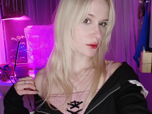 MiraliXSweet with blonde hair, pink lipstick, and a nose ring, wearing a black jacket over a pink top, holding her jacket's edge, in a purple-lit room.