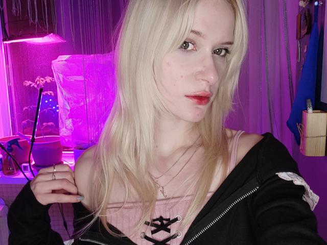 MiraliXSweet with blonde hair, pale skin, and red lipstick, wearing a pink lace-up top and black jacket, holding a ring, in a purple-lit room.