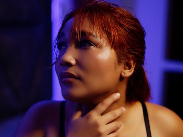 MiiaGrey with wet red hair, brown skin, and subtle makeup, touches her neck while gazing to the side under blue lighting, showing smooth, shiny skin and black tank top.