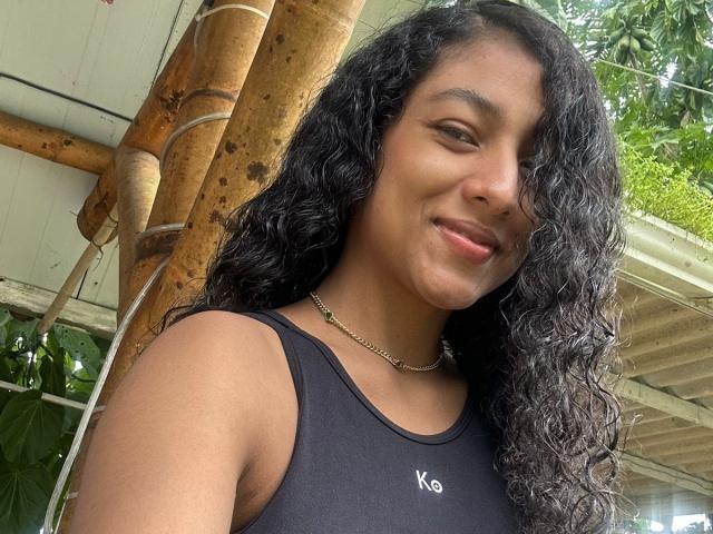 NatalyBlonder with curly black hair, medium brown skin, smiling, wearing a black K-Swim tank top, and a gold chain necklace, standing next to a bamboo pole.