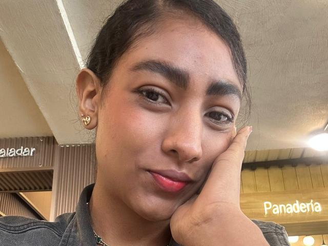 NatalyBlonder's face, brown skin, black hair in a ponytail, small hoop earrings, pink lipstick, hand resting on cheek, wearing a gray jacket, subtle smile, natural makeup.