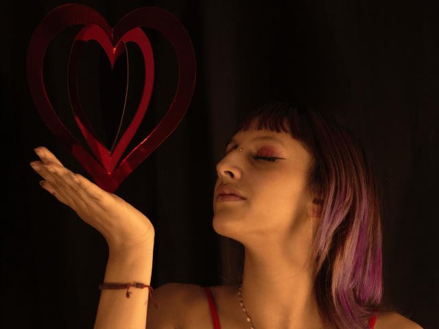 KatsuraRen has short, straight purple and brown hair with bangs. She's wearing a red spaghetti strap top and a simple bracelet. She's holding a red heart-shaped object above her right hand, eyes closed and lips slightly parted. Her makeup includes pink eyeshadow. The dark background contrasts with her light skin and the red heart. The lighting is warm, highlighting her face and the heart. Her expression is calm and focused. The composition is centered on KatsuraRen and the heart.