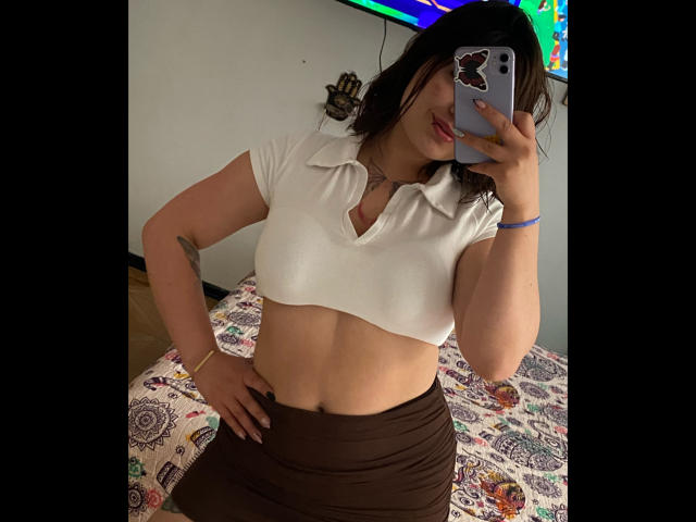 AnikaFleur in a white cropped shirt showing her tits, brown skirt, holding a phone with a cat case, taking a mirror selfie, her hand on her hip, dark hair.