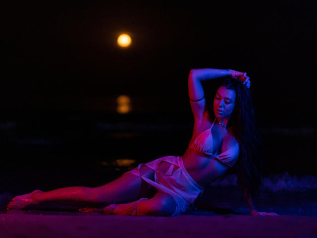 NatashaDavie is laying on a beach at night, wearing a bikini, with her arm above her head, glowing in blue and red lights, showing her tits and ass.