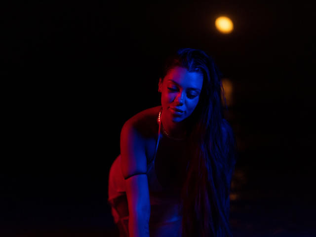 NatashaDavie with blue and red lighting, leaning forward, long wet hair, bare shoulders, necklace, looking down, dark background, single orange light, moody and sexy, night scene.