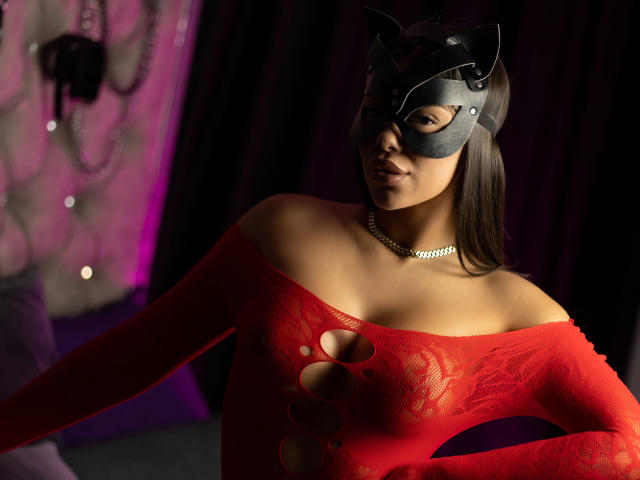  NatashaDavie wears a black cat mask with pointed ears, a red lace off-shoulder bodysuit with cut-out sections, and a silver chain necklace. Her dark hair flows past her shoulders. The bodysuit's texture is detailed with floral patterns. She's in a dimly lit room with purple and black curtains. Her expression is calm, slightly smiling. The word "cosygirl" is visible on the chain necklace. The overall style is sexy and provocative.