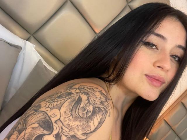 AnnyBentleii tiene el cabello largo y negro, piel clara y un tatuaje detallado de un dragón en el hombro derecho. Está mirando a la cámara con lápiz labial rosa..
