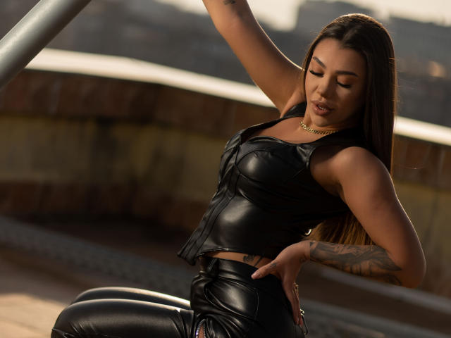 NatashaDavie with long brown hair, wearing a black leather crop top and matching pants, has a tattoo on her right arm, gold necklace, and is leaning against a metal rail.