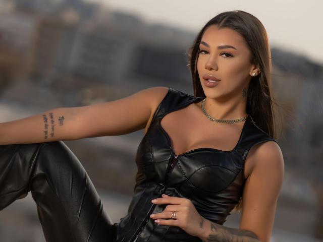 NatashaDavie with long brown hair, wearing black leather top and pants, has tattoos on arms, chain necklace, and ring, sitting with legs up, showing small boobs and confident smile.