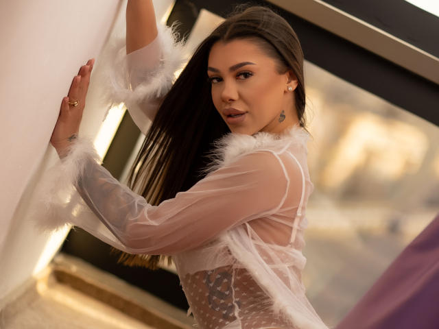 NatashaDavie stands against a white wall, looking back over her shoulder. She has long brown hair and wears a sheer white top with feathered sleeves and a polka dot pattern. Her right hand is placed on the wall, and her left arm is slightly raised. She has a small tattoo on her right shoulder. The light is soft, creating a warm glow on her skin. She has a confident, slightly seductive expression. The image has a slightly blurred background with a large window. The composition emphasizes her upper body and facial expression.