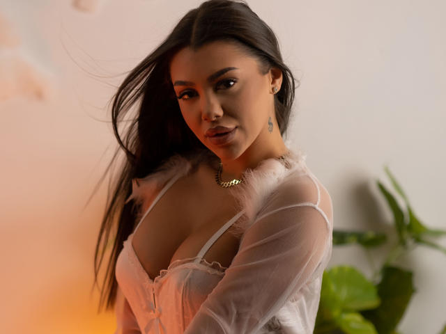 NatashaDavie with dark hair, wearing a sheer white top with fluffy edges, showing her big tits, looking at the camera, smiling, gold necklace, small tattoo on her neck.