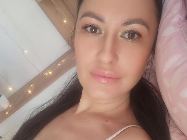 O rosto de Frederica, com lábios rosados e brilhantes, olhos castanhos e longos cabelos pretos, vestindo uma alça branca, olhando para a câmera com um leve sorriso. Luzes de fada suaves ao fundo..
