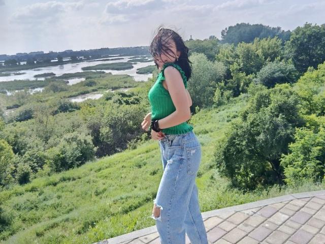 SweetieChloe with medium tits in a green sleeveless top and ripped blue jeans, standing on a brick patio, greenery and water in the background, wind in her dark hair.