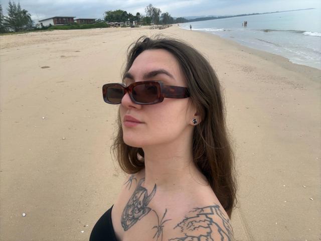 A photo of MilaSusse-hot on a beach. She's facing slightly to the right, wearing brown tortoiseshell sunglasses and a black strapless top. She has long brown hair, light skin, and several tattoos on her chest and shoulder, including a detailed dragon. Her lips are slightly parted, and she's wearing small black earrings. The beach has a wide, sandy shoreline with sparse greenery and a few buildings in the distance. The ocean is to the right with gentle waves. The sky is overcast.