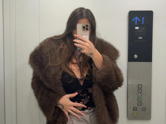 MilaSusse-hot takes a mirror selfie in an elevator. She wears a fluffy brown fur coat over a black lace top with deep cleavage, showing a tattoo on her right hand. Her long brown hair is loose, and she holds an iPhone with a gold case, covering her face. Her right hand is on her belly, and she has light skin and pink nails. The elevator button panel is on the right, with a blue arrow indicating the elevator is moving. The image is clear and modern.