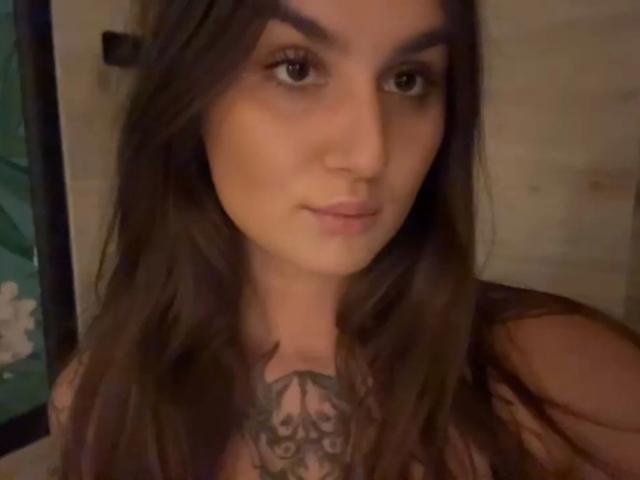 MilaSusse-hot has long brown hair, a tattoo on her chest, and a neutral expression. She's looking at the camera with brown eyes and slightly parted lips.