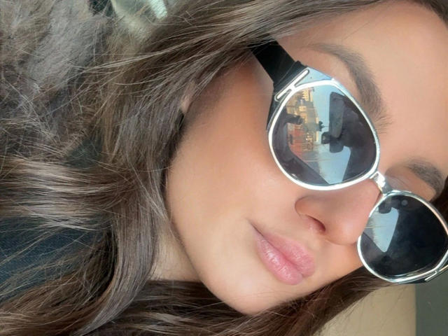 MilaSusse-hot with glossy brown hair, wearing reflective aviator sunglasses, showing a reflection of a cityscape in her mirrored lenses, with pinkish lips and a subtle smile.