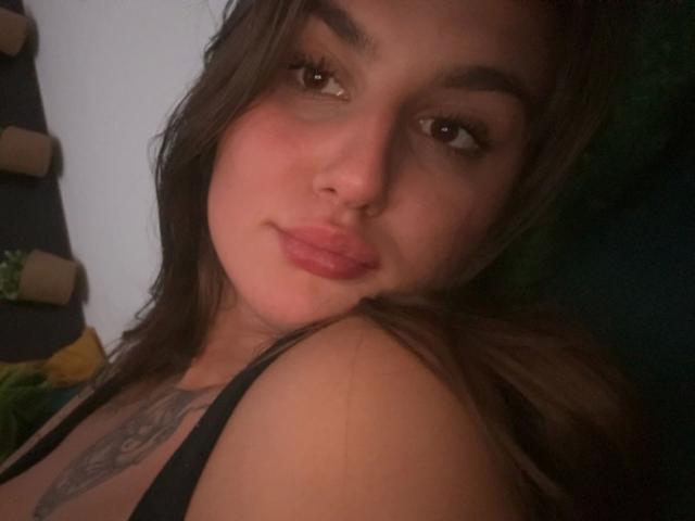 MilaSusse-hot with pouty lips, brown eyes, and long brown hair, wearing a black top, showing a tattoo on her chest, looking at the camera.