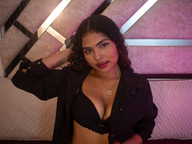 AshlyCorines with curly black hair, medium brown skin, wearing a black bra and unbuttoned shirt, hand in hair, red lipstick, gold necklace, showing her tits, standing in a room.