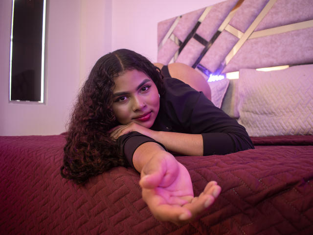 AshlyCorines with curly black hair, lying on a red quilted bed, wearing a black shirt, reaching out with her right hand, looking at the camera with pink lipstick.