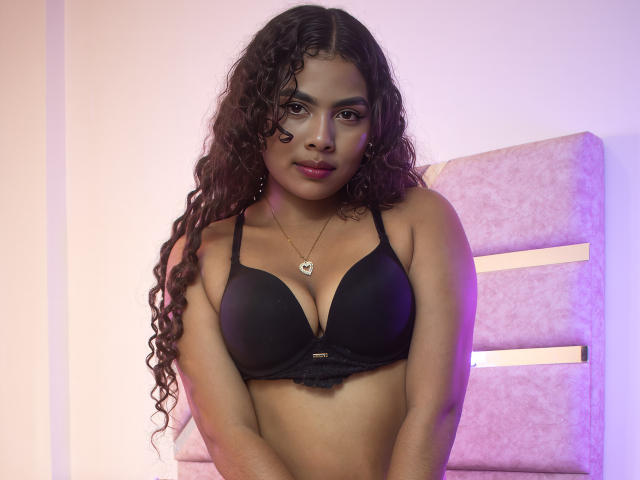 AshlyCorines with long curly brown hair, dark skin, wearing a black bra, showing her tits, a small necklace, and a subtle purple light in the background.