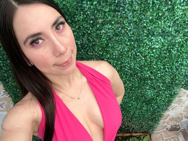 FreyaraVenwo with long brown hair, pink eyeliner, and pink halter top, showing cleavage, smiling at camera, green leafy wall in background, gold necklace, cute, attractive.