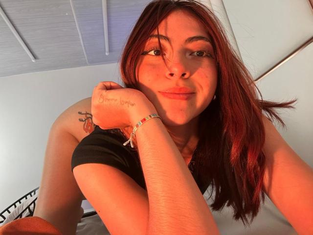 SpencerLe with red hair, smiling, black top, tattoo on right buttcheek, arm tattoo, bracelet, leaning forward, looking at camera, orange lighting, white ceiling, wooden beams.