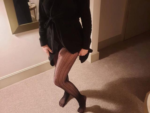 HotSexyPaar-hot wearing a black dress, standing with legs crossed, black patterned tights, hands on dress, in a carpeted room, shadow on floor, mirror on left wall.