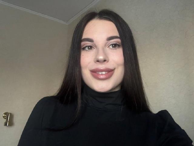 A close-up photo of BlackFatal. She has long, straight black hair and is wearing a black turtleneck. Her eyebrows are dark and well-defined, and she has light pink eyeshadow. She's smiling slightly with glossy, pinkish lips. The background is a plain, beige wall with a small, gold door handle on the left. The image is centered on her face, which is lit evenly. She has a fair skin tone and a natural, attractive look. Her eyes are dark brown, and she has a subtle, confident expression.