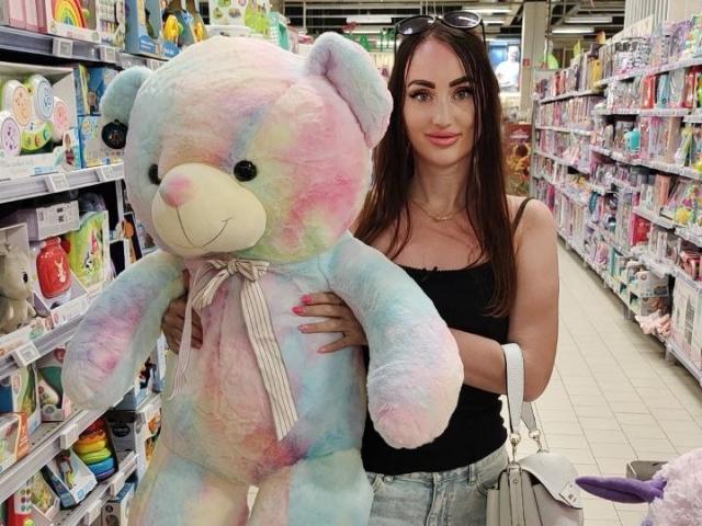 FortuneLisa6 holds a large, pastel-colored stuffed teddy bear with rainbow hues and a white ribbon. She has long brown hair, light skin, and wears a black tank top and denim shorts. Her right hand is on the bear's back, left arm around its side. Shelves of similar toys and plushies are in the background. She has a slight smile, eyeglasses on her head, and a white handbag. The image is bright and colorful.