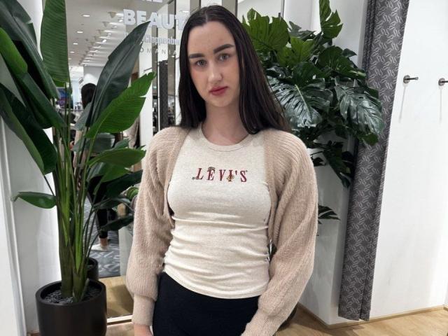FortuneLisa69, met lang bruin haar, een beige vest, een wit Levi's shirt en zwarte broek, staat in een winkel, heeft een gemiddelde borstmaat en een schattig gezicht, en kijkt naar de camera..