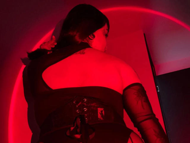 Jesskhy is in a dark, red-lit room with a circular light source behind her. She has dark hair in a ponytail, wearing a black, strapless top with a large cutout, and a lace-up corset. Her right arm is covered in a black, semi-transparent sleeve with a tattoo visible on her upper arm. The red light casts dramatic shadows, highlighting her back and the corset's details. The composition focuses on the contrast between the dark clothing and the red light.