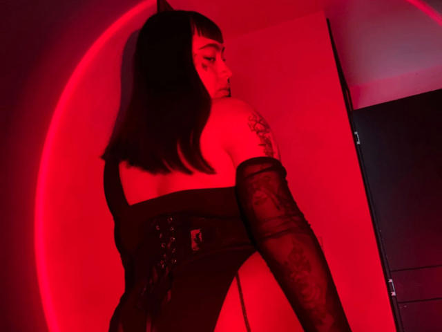 Jesskhy with red-lit skin, black strapless dress, corset, lace arm sleeves, and cat ears, showing a tattoo on her left shoulder, looking back. Red glow highlights her.