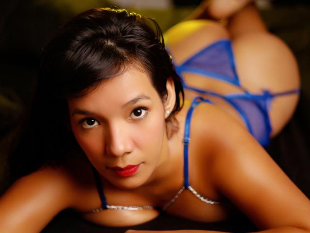 LuciaMoretti with dark hair, red lipstick, blue lingerie, lying on her stomach, showing her ass and looking at the camera with a slightly open mouth.